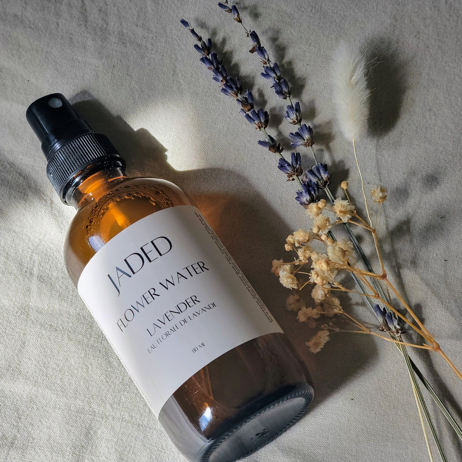 A brown glass bottle labeled 'JADED' with 'FLOWER WATER' and 'LAVENDER' above it, alongside dried lavender flowers and foliage on a light background.