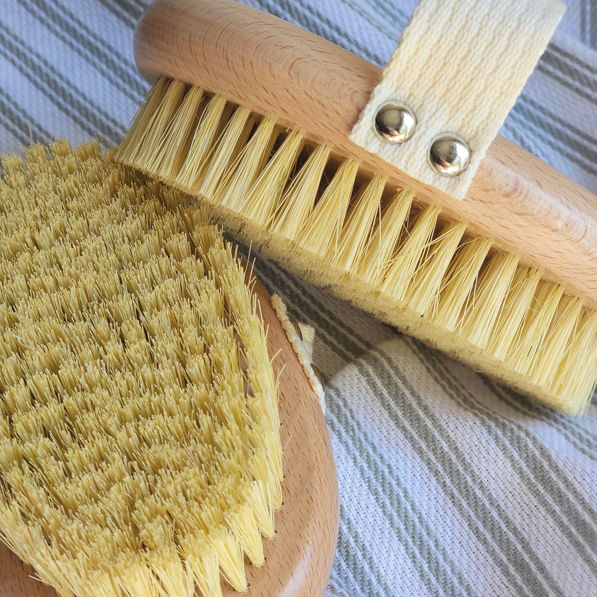 A close-up of a beechwood and sisal bristle dry brush with a cotton hand strap, laid out on a striped towel.