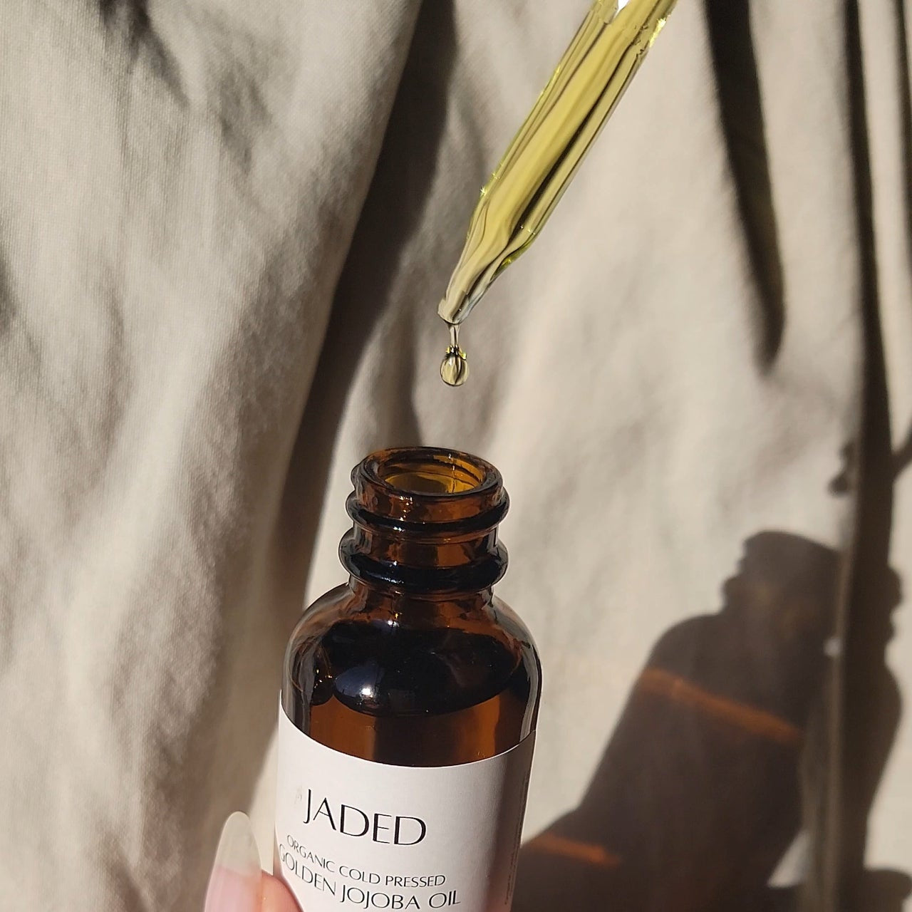 Hand holding a bottle of Jaded jojoba oil with a dropper, against a neutral background.